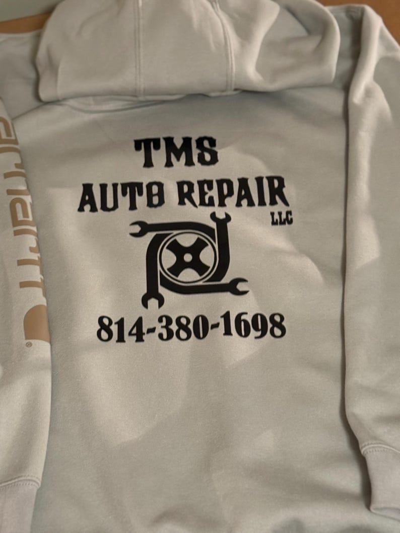 K&ouml;nnte beinhalten: Hellgrauer Kapuzenpullover mit dem Text "TMS AUTO REPAIR LLC" in schwarzen Blockbuchstaben. Darunter befindet sich eine Grafik eines Reifens und Schraubenschl&uuml;sseln. Die Telefonnummer 814-380-1698 ist darunter aufgedruckt. Das Carhartt-Logo ist auf dem &Auml;rmel zu sehen.