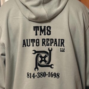 K&ouml;nnte beinhalten: Ein hellgrauer Kapuzenpullover mit dem schwarzen Text "TMS AUTO REPAIR LLC". Das Design umfasst eine Grafik eines Reifens und Schraubenschl&uuml;sseln sowie die Telefonnummer 814-380-1698.