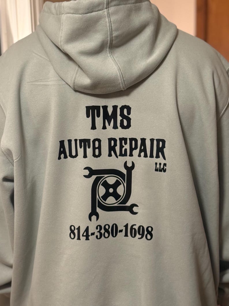 K&ouml;nnte beinhalten: Ein hellgrauer Kapuzenpullover mit dem schwarzen Text "TMS AUTO REPAIR LLC". Das Design umfasst eine Grafik eines Reifens und Schraubenschl&uuml;sseln sowie die Telefonnummer 814-380-1698.