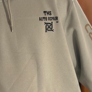 K&ouml;nnte beinhalten: Hellgr&uuml;ner Kapuzenpullover mit Fronttasche und Kordelzugkapuze. Der Text "TMS AUTO REPAIR LLC" ist in Schwarz auf der Brust aufgedruckt, zusammen mit einer Grafik aus Schraubenschl&uuml;ssel und Zahnrad. Das Carhartt-Logo ist auf der Tasche zu sehen.