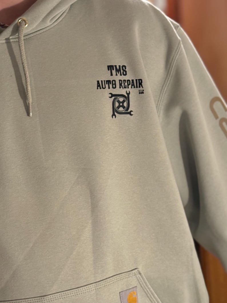 K&ouml;nnte beinhalten: Hellgr&uuml;ner Kapuzenpullover mit Fronttasche und Kordelzugkapuze. Der Text "TMS AUTO REPAIR LLC" ist in Schwarz auf der Brust aufgedruckt, zusammen mit einer Grafik aus Schraubenschl&uuml;ssel und Zahnrad. Das Carhartt-Logo ist auf der Tasche zu sehen.