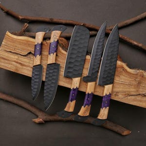 May include: A set of five handcrafted kitchen knives with black blades and wooden handles. The handles feature a purple accent and are set on a wooden base. The blades have a textured surface.