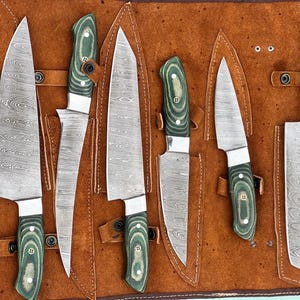 May include: A set of seven kitchen knives with patterned steel blades and green wooden handles, displayed in a brown leather knife roll. The blades vary in size and shape, suitable for various culinary tasks.