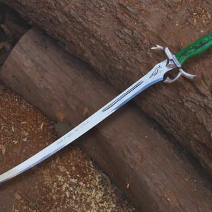 May include: A curved sword with a silver blade and a green handle with a decorative design. The sword has a silver hilt with curved accents. The blade has text on it. The sword is resting on a wooden surface.
