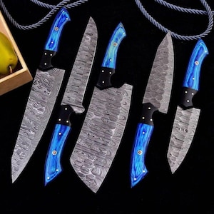 May include: Set of six Damascus steel kitchen knives with blue wooden handles. The blades have a unique patterned design. The handles are a vibrant blue with black accents and brass rivets. Knives are arranged on a black surface.