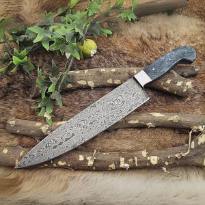 May include: A Damascus steel chef's knife with a dark gray handle and a silver bolster. The blade features a unique swirling pattern. The knife is displayed on a bed of twigs and fur, with green foliage in the background.