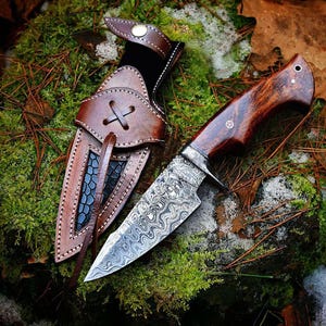 May include: A handcrafted hunting knife with a patterned steel blade and a dark wood handle. The knife is displayed with a brown leather sheath, featuring decorative stitching and a cross-shaped closure. The knife and sheath are set against a backdrop of moss and leaves.