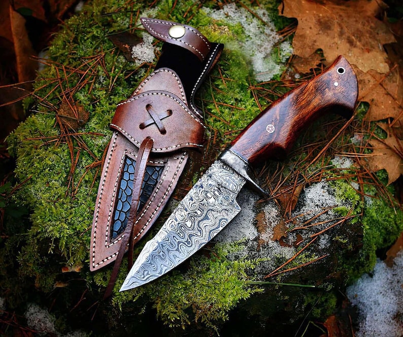 May include: A handcrafted hunting knife with a patterned steel blade and a dark wood handle. The knife is displayed with a brown leather sheath, featuring decorative stitching and a cross-shaped closure. The knife and sheath are set against a backdrop of moss and leaves.