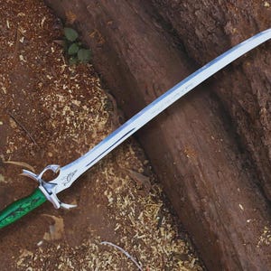 May include: A silver fantasy sword with a curved blade and a green handle. The sword has intricate details on the hilt and blade, and is lying on a wooden surface.