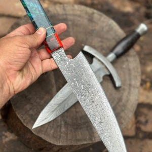 May include: A handmade chef's knife with a patterned blade and a colorful handle. The handle features a combination of red, blue, and green resin. A second knife with a dark wooden handle and a silver blade is in the background.