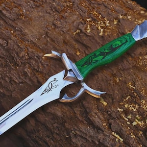 May include: A fantasy-style sword with a silver blade and a green wooden handle. The handle has a dragon design etched into it. The sword rests on a brown, textured surface, possibly wood.