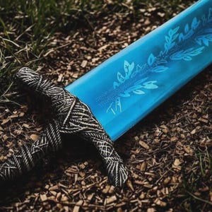 Könnte beinhalten: Ein Fantasy-Schwert mit einer leuchtend blauen Klinge mit einem Blattdesign. Der Griff ist aufwendig in einem schwarz-weißen Muster gewickelt. Das Schwert liegt auf einem Bett aus Holzspänen und Gras.