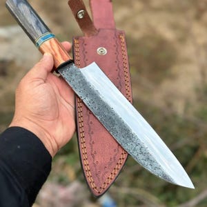 May include: A handcrafted chef's knife with a textured blade and a wooden handle with blue accents. The knife is paired with a brown leather sheath. The blade is approximately 12 inches long.