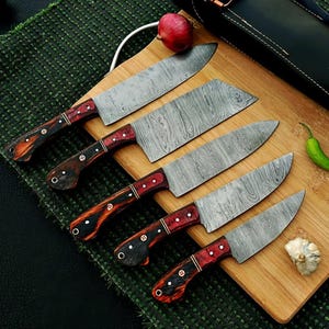 May include: A set of five Damascus steel kitchen knives with wooden handles. The handles are a mix of red and brown tones, with a dark wood grain pattern. The blades have a unique, textured pattern. The knives are arranged on a wooden cutting board.