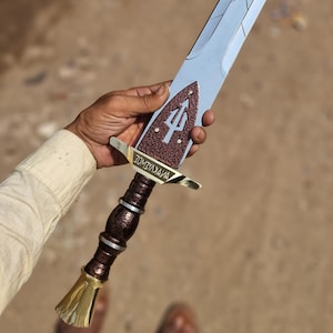 Op de afbeelding: Een decoratief zwaard met een lichtblauw lemmet en een bruin handvat. Het handvat heeft een goudkleurige pommel en een bruine grip. Het zwaard heeft een bruine en gouden pareerstang en een bruin gedeelte met een drietandsymbool. De tekst "ZOMYAXAVA" is zichtbaar.