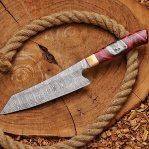 May include: A Damascus steel chef's knife with a red, black, and white handle. The blade has a unique wood grain pattern. The knife is set on a wooden surface with a rope border.