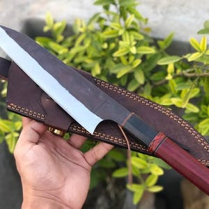 May include: A large, hand-forged chef's knife with a dark blade and a polished edge, resting on a brown leather sheath. The handle is a dark wood with a red end. The knife is approximately 14 inches long.