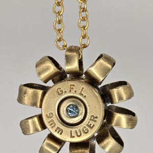 May include: A gold-colored pendant necklace featuring a repurposed 9mm Luger bullet casing. The casing is adorned with a light blue gemstone and surrounded by stylized petal-like loops. The chain is also gold-colored.