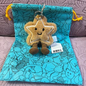 Peut inclure: Un porte-clés en forme d'étoile en peluche avec un visage souriant et des jambes marron, attaché à un porte-clés argenté. Il repose sur un sac à cordon de serrage bleu sarcelle orné de motifs d'ours de dessin animé. Le sac a des cordons jaunes.