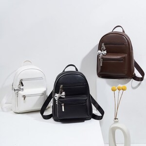 May include: Three small backpacks in white, black, and dark brown. Each bag features a top handle, adjustable shoulder straps, two front zippered compartments, and a silver-toned bow charm attached to the upper zipper pull.