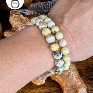 Op de afbeelding: Een dubbelstrengs armband gemaakt van crèmekleurige, ronde kralen met gele en grijze accenten. De kralen zijn aan elkaar geregen, waardoor een textuur en natuurlijk uiterlijk ontstaat. De armband wordt om een pols getoond.