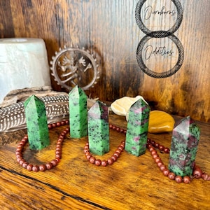 Puede incluir: Cinco torres de cristal de rubí zoisita verdes y rojas se exhiben sobre una superficie de madera, conectadas por un hilo de cuentas rojas. La imagen también incluye el texto "Ouroboros Oddities".