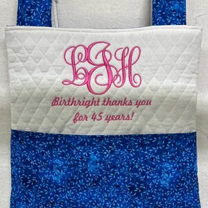 May include: A quilted tote bag with blue floral patterned fabric at the bottom and top handles. The center is white with a pink embroidered monogram and the text "Birthright thanks you for 45 years!"