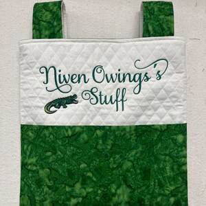 May include: A white quilted hanging organizer with green fabric straps and a green fabric bottom. The organizer has an embroidered alligator and the text "Niven Owings's Stuff" in green thread.