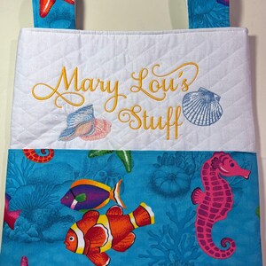 May include: A quilted white and blue fabric tote bag with the embroidered text "Mary Lou's Stuff". The bag features a colorful aquatic design with fish, a seahorse, and starfish. The top has two blue fabric handles.