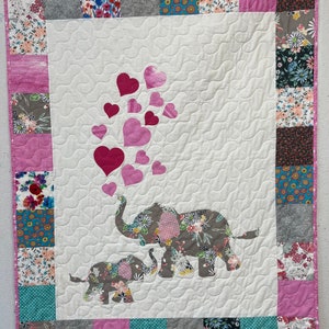 May include: A white quilt with a pink border and a floral pattern. The quilt features two gray elephants with floral patterns, one large and one small, with pink hearts floating above them.