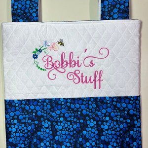 May include: White quilted fabric storage bag with blue polka dot fabric handles and bottom. The bag has a pink embroidered design that says "Bobbi's Stuff" with a bee and flowers.