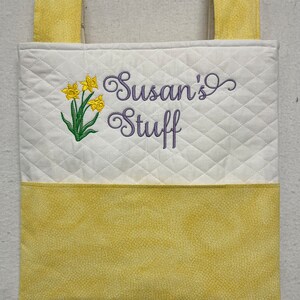 May include: A white quilted hanging organizer with yellow fabric straps and a yellow fabric bottom. The organizer has an embroidered design of yellow daffodils and the text "Susan's Stuff" in purple thread.