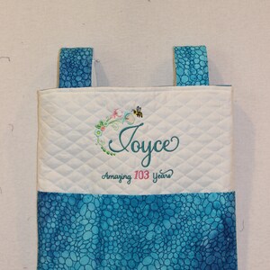 May include: A white quilted tote bag with blue fabric handles and a blue and teal patterned bottom. The bag has an embroidered design that says "Joyce Amazing 103 Years" with a bee and flowers.