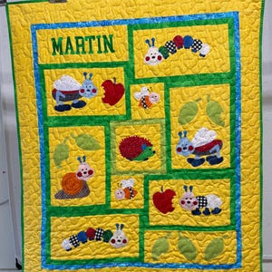May include: A handmade yellow baby quilt with a blue and green border. The quilt features appliqued images of insects, fruit, and leaves. The name "MARTIN" is embroidered in green letters at the top.