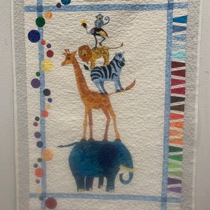 May include: A handmade quilt featuring a whimsical design of stacked animals. A blue elephant supports a giraffe, lion, zebra, and other creatures. The quilt has a white background with colorful accents and a blue border.