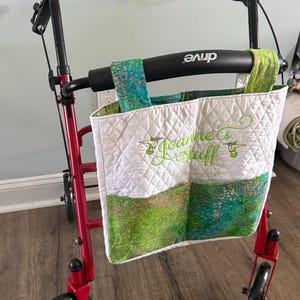 May include: A red four-wheeled walker with a black frame and handle, featuring a white quilted fabric storage bag attached to the front. The bag has green and blue patterned pockets and is embroidered with the text Joanne's Stuff and hummingbirds.
