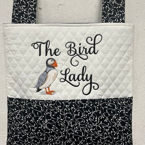 May include: A white quilted tote bag with black fabric handles. The bag has a black and white patterned fabric lining. The bag is embroidered with the text "The Bird Lady" and an image of a puffin.