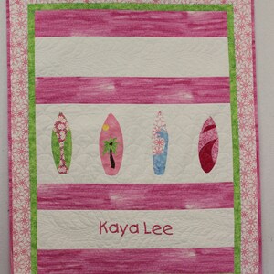 May include: A pink and white striped quilt with a green border and four surfboards. The quilt is personalized with the name "Kaya Lee".