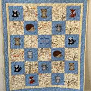 May include: A blue and cream baby quilt with a checkered pattern. Each square features an embroidered woodland animal, including a bear, fox, raccoon, owl, hedgehog, and skunk. The quilt is personalized with the name "Asa Chapin" and the date "March 5, 2022".