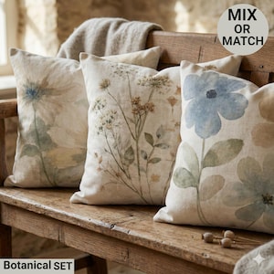 Op de afbeelding: Vier decoratieve sierkussens met botanische ontwerpen liggen op een houten bank. De kussens hebben aquarelprints met bloemen in blauw, beige en groen. De tekst "MIX OR MATCH" staat in een cirkel rechtsboven. De tekst "Botanical SET" staat onderaan.