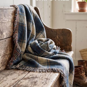 May include: A woven throw blanket featuring a blue, cream, and brown plaid pattern with fringed edges. The blanket is draped over a wooden bench. The text Woven Patina Blue Plaid is visible at the bottom, and the text SHIPS FREE is at the top.