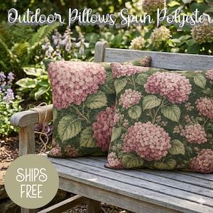 May include: Two outdoor pillows with a floral hydrangea design in shades of pink, green, and white. The pillows are made of spun polyester and rest on a wooden bench. The text "Outdoor Pillows Spun Polyester" is at the top.