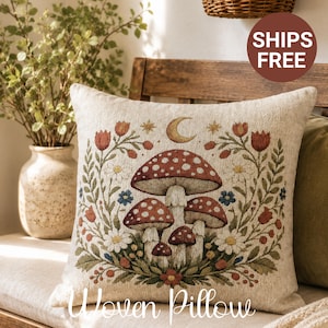 May include: A square throw pillow with a beige textured fabric base featuring a central woven design of red and white mushrooms surrounded by a floral wreath of red tulips, white daisies, and blue flowers, topped with a golden crescent moon and stars.