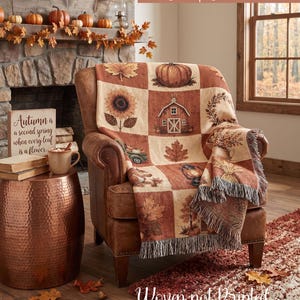 May include: A woven throw blanket featuring a grid pattern with autumn-themed illustrations including pumpkins, a sunflower, a barn, a wreath, leaves, a lantern, and a truck. The blanket has a fringed edge and displays warm orange, brown, and cream tones.