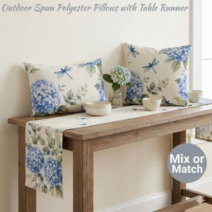 Può includere: Due cuscini da esterno in poliestere filato e un runner da tavolo con un motivo floreale di ortensie e libellule. I cuscini e il runner sono color crema con accenti blu e verdi. Due piccole ciotole bianche e un vaso di fiori sono sul tavolo di legno.