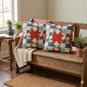 May include: Two square throw pillows with a patchwork star design in red, blue, and cream colors. The pillows are placed on a wooden bench. The bench is in a room with a window and plants.