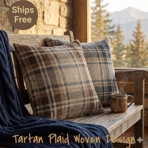May include: Two square plaid cushions in brown, blue, and tan tones sit on a wooden bench with a blue knitted throw. A ceramic mug is also present. The cushions have a tartan plaid woven design.