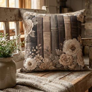 May include: Square decorative pillow featuring a print of antique books and floral accents in shades of brown, beige, and cream. The pillow is placed on a rustic wooden bench, with a small plant in a ceramic pot visible in the foreground.
