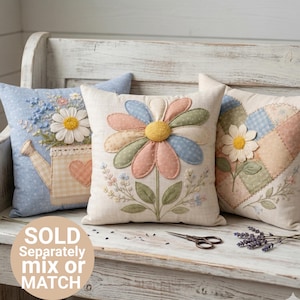 May include: Three decorative throw pillows with floral designs. One pillow features a large daisy, another has a watering can and daisy, and the third has a patchwork design with a daisy. The pillows are on a white bench.