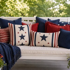 Op de afbeelding: Een verzameling kussens en een deken met franjes in rood, marineblauw en crème op een witte schommel. De kussens hebben sterpatronen. De scène speelt zich buiten af met een achtergrond van groen gebladerte, wat een ontspannen buitenomgeving creëert.